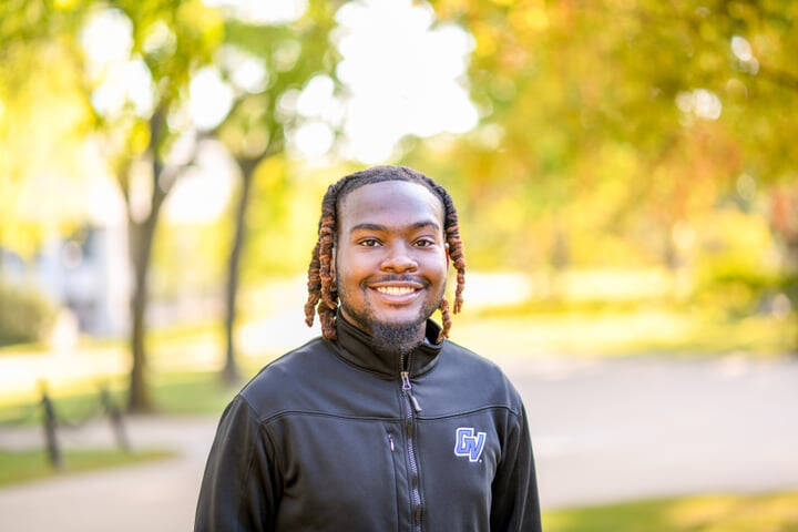 Dogan Copeland stands smiling in front of trees on a sunny day. Dogan is wearing black GVSU spirit wear and has black and brown hair.